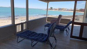 A covered balcony with two reclining chairs and a table, overlooking a beach with clear blue water and a distant shoreline.