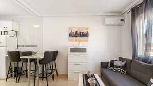 A modern living room with a grey sofa, a dining area with two chairs, a white coffee table with decorative items, a white cabinet, and a cityscape painting on the wall. - Vacation home Hamber by Interhome (Puerto de Mazarrón)