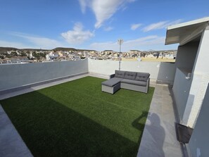 A rooftop terrace with artificial grass, a grey sectional sofa, and a clear view of a coastal town.