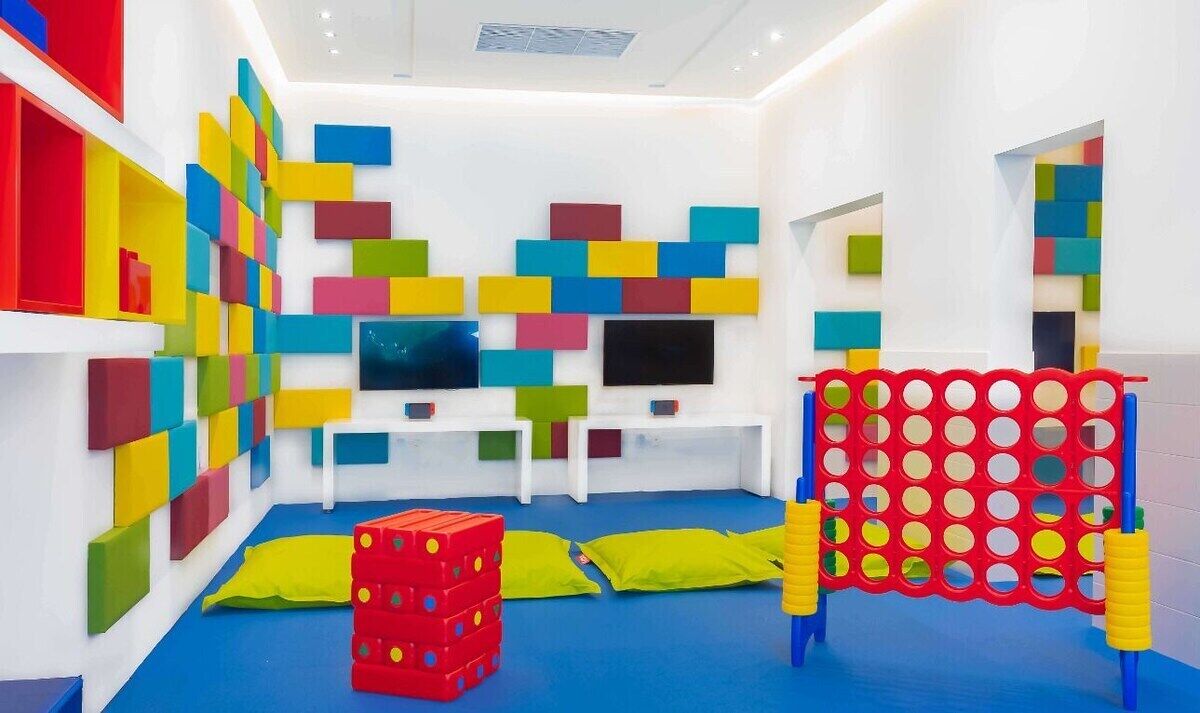 Uma sala de brinquedos com blocos coloridos, uma TV e um cercadinho vermelho e amarelo.