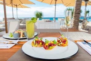 Una mesa dispuesta con tacos, una copa de vino blanco y un cóctel en un patio frente a la playa, con sombrillas y vista al océano.