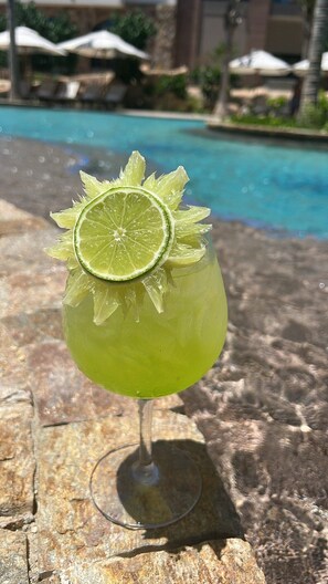 Een cocktail met een limoenpartje in een glas, geplaatst op een stenen oppervlak nabij een zwembad.