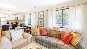 A living room with a sofa, a coffee table, and a dining area with chairs and a table. There are two windows with curtains, a kitchen area with cabinets and a microwave, and a small table with a plant. - A Walk From the Beach, 2bed 2bath apartment in 3unit building 1 block from Ocean (North Myrtle Beach)