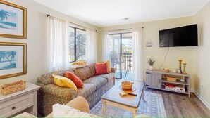 A living room with a sofa, coffee table, and a television. There is a balcony with a view of buildings. - A Walk From the Beach, 2bed 2bath apartment in 3unit building 1 block from Ocean (North Myrtle Beach)