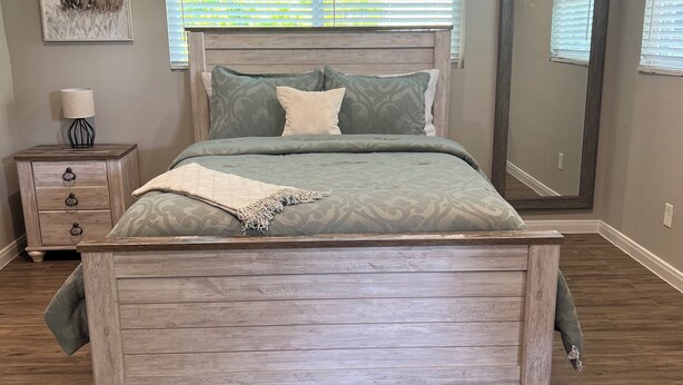 Une chambre à coucher avec un lit en bois, une table de chevet, une lampe, un tableau encadré et une fenêtre avec des stores.