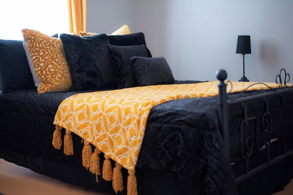 Una cama con estructura negra, una colcha amarilla estampada y varias almohadas en tonos negro y amarillo.