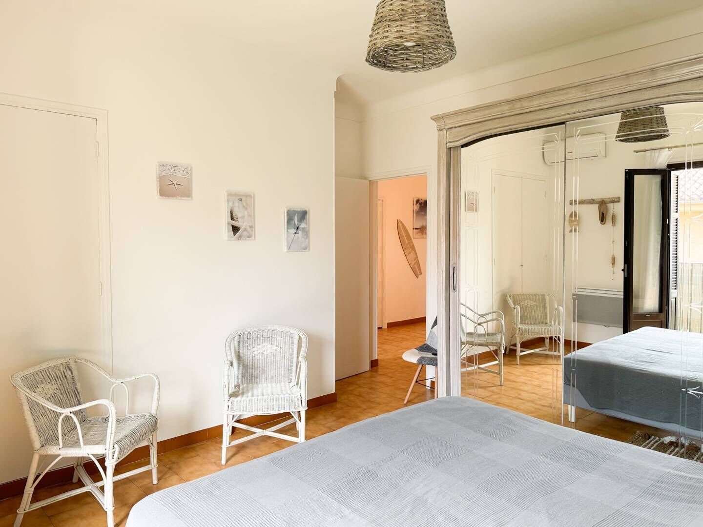 Una habitación con una cama grande, dos sillas blancas de mimbre, una lámpara de mimbre y vistas a una playa a través de un ventanal.