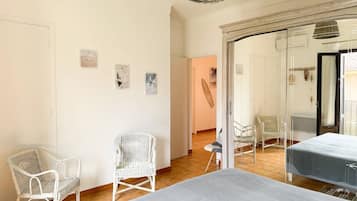Una habitación con una cama grande, dos sillas blancas de mimbre, una lámpara de mimbre y vistas a una playa a través de un ventanal.