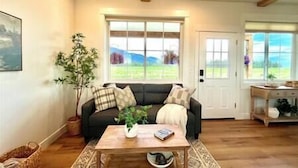 A living room with a sofa, coffee table, and a large window offering a view of the outdoors. - Luxe 1br Cottage - Mtn Views! "Cedar" (Columbia Falls)