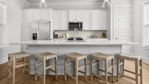 Una cocina moderna con armarios blancos, una isla central con taburetes, electrodomésticos de acero inoxidable e iluminación colgante.