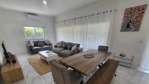 A modern living room with a large wooden dining table, grey sofas, and a TV on a stand.