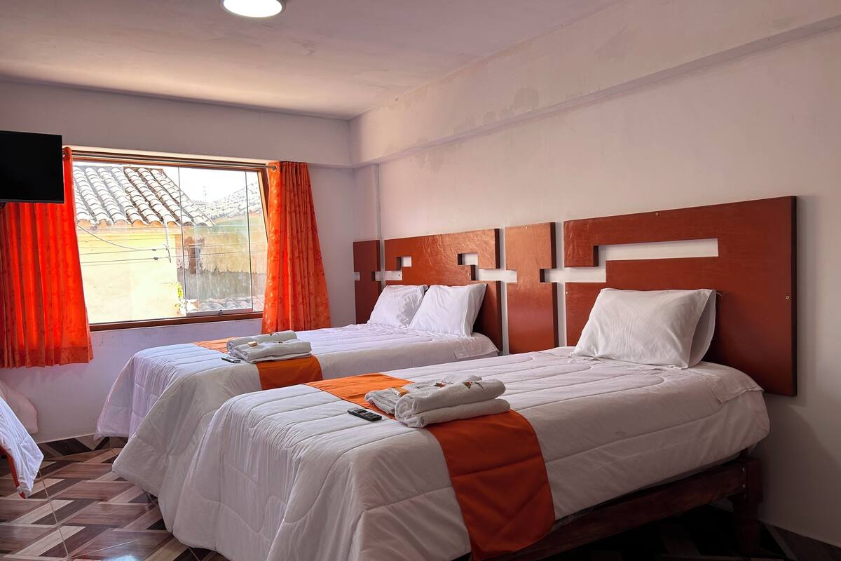 Habitación de hotel con dos camas, ventana con cortinas naranjas, televisor de pantalla plana y piso de baldosas.