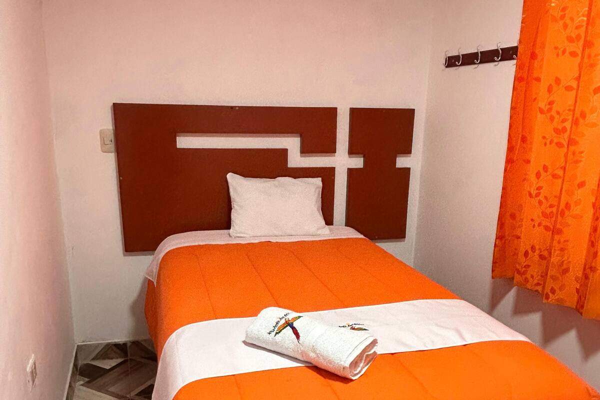 Una cama individual con colcha naranja y almohada blanca, cabecera de madera y cortina naranja estampada.