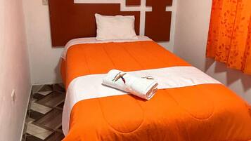 A single bed with an orange bedspread and a white pillow, a wooden headboard, and a patterned orange curtain.