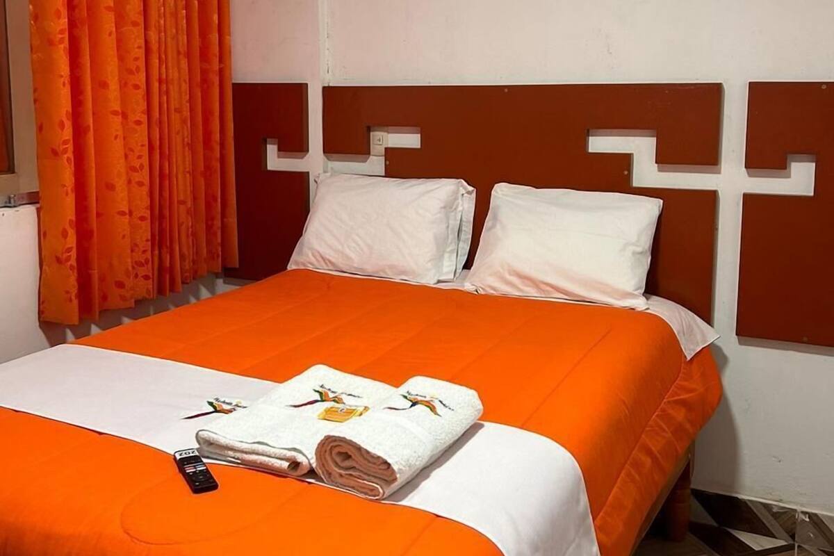 Una habitación de hotel con cama, cortinas naranjas, almohadas blancas y cabecero estampado.