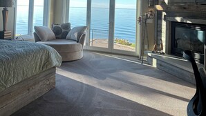 A bedroom with a large bed, a ceiling fan, a fireplace, a TV, and a view of the ocean through large windows.