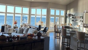 A spacious living area with a large window overlooking the ocean, a kitchen with a bar, and a ceiling fan.