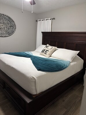 Un lit bien fait, avec un matelas blanc, un plaid turquoise et deux oreillers, dont l’un porte l’inscription « HOME ».