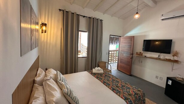 A hotel room with a bed, a TV, a window with curtains, and a wooden ceiling. - AROEIRA POUSADA TRANCOSO (Porto Seguro)