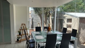 A modern dining area with a glass table and black chairs, a wooden sideboard with plants, and a view of a stone building and trees outside. - 3 Bedroom cozy villa in Werribee near  Werribee CBD and other amenities (Werribee)
