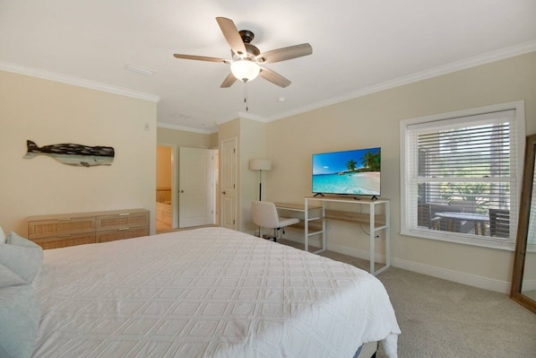 A bedroom with a bed, a desk with a computer, a ceiling fan, a TV displaying a beach scene, and a window with blinds.