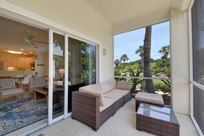 A balcony with a wicker sofa, a glass-top table, and a view of a tropical landscape.