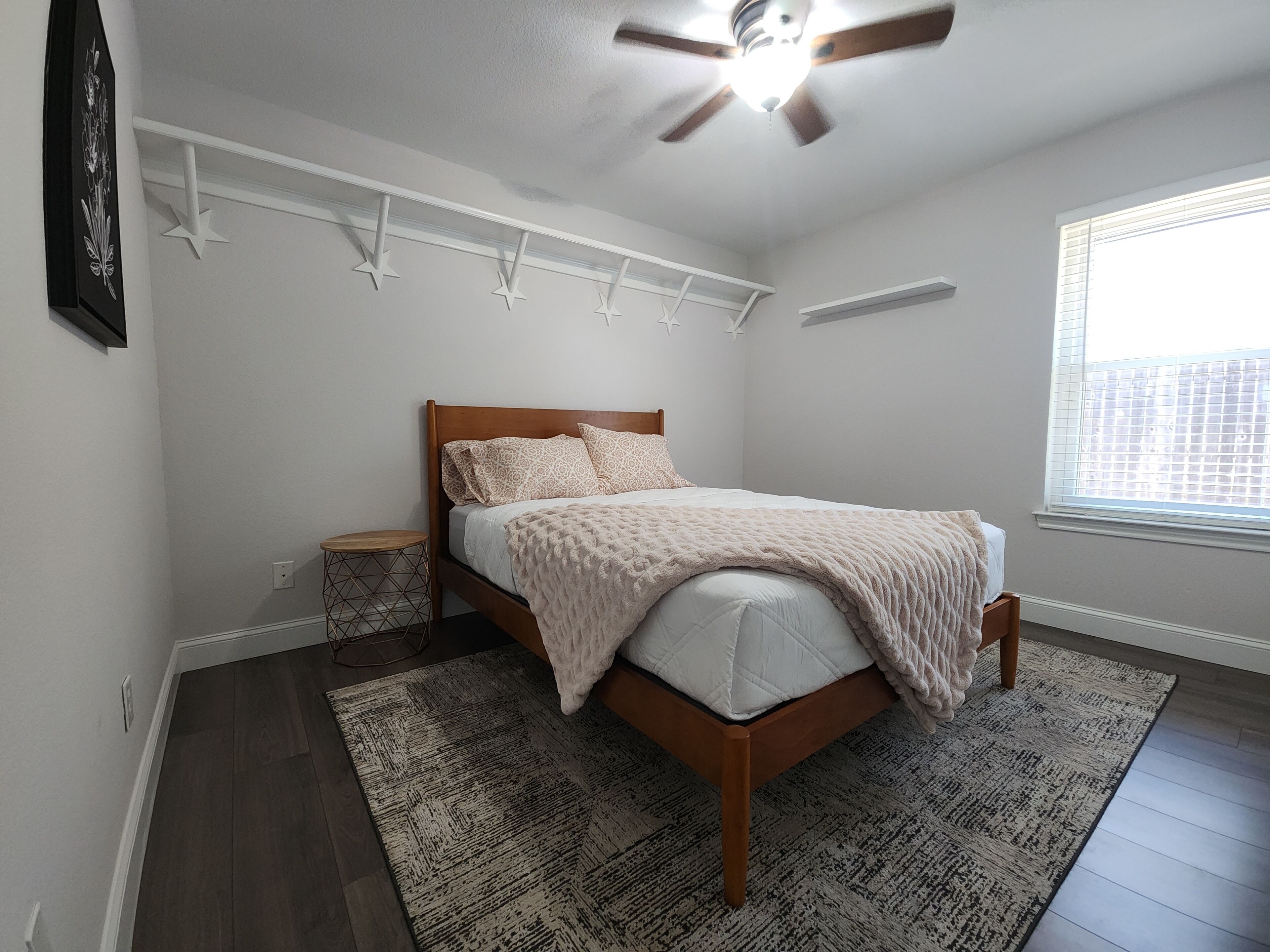 A bedroom with a wooden bed, white walls, a ceiling fan, a window with blinds, and a patterned rug.