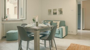 A modern living room with a dining table set for two, a sofa, and a window with a view of a building. - Diana - MyHomeinComo (Como)