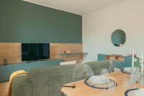 A modern living room with a sofa, a dining table set for two, a TV mounted on a wooden panel, and a small shelf with decorative items.