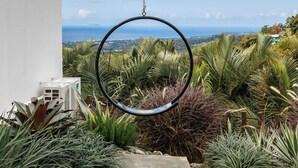 A hanging ring chair suspended from a balcony railing overlooking a garden with various plants and a view of the ocean. - Adults Only! Desecheo Suite II at Ocaso Luxury Villas (Rincon)