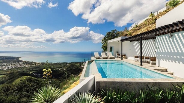 A modern pool with a view of the sea, surrounded by lush greenery and a white building with a pergola. - Adults Only! Desecheo Suite II at Ocaso Luxury Villas (Rincon)