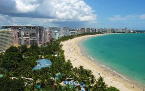 Paisagem costeira com uma extensa praia de areia, mar azul cristalino e edifícios altos ao fundo.