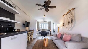 A modern hotel room with a kitchenette, a dining area, a living room with a sofa, a ceiling fan, and a macrame wall hanging.