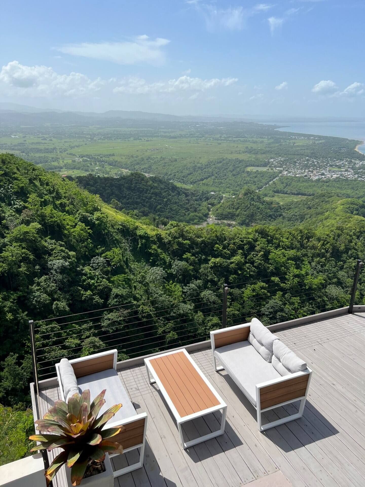 A deck with a sofa and a table, overlooking a lush green valley and a distant town.