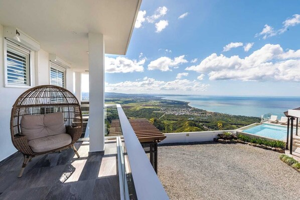 Un balcon avec un fauteuil en osier, une piscine et une vue panoramique sur la mer et le paysage.