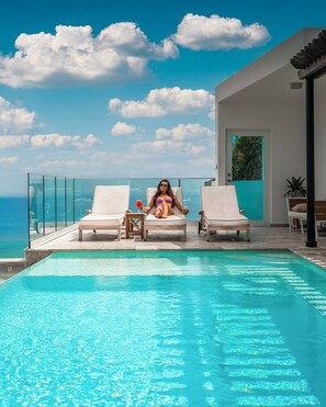 A poolside area with a clear view of the ocean, lounge chairs, and a modern building with a glass door.