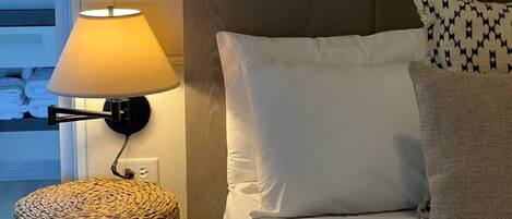 A neatly made bed with white and patterned pillows, a woven side table, and a wall-mounted lamp.