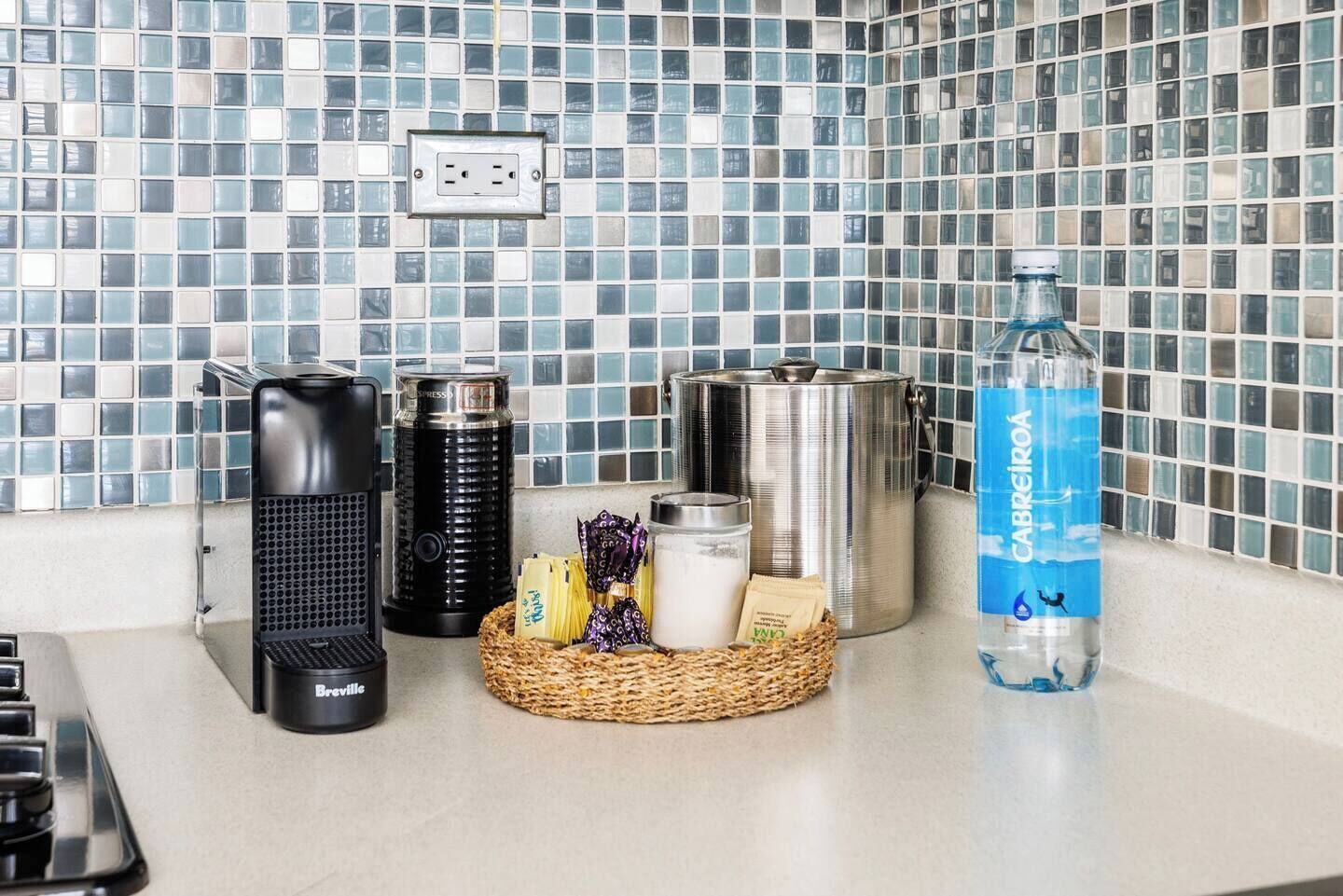 Un comptoir de cuisine avec une machine à café, un distributeur d’eau, une bouilloire, un thermos et un panier rempli d’en-cas.