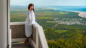 A person in a white robe standing on a balcony with a view of a coastal landscape.