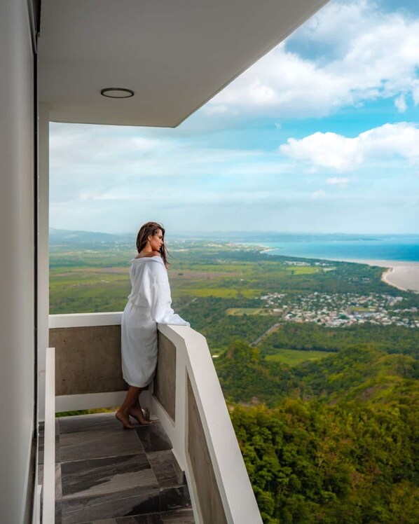 A person in a white robe standing on a balcony with a view of a coastal landscape.