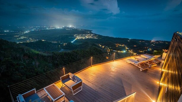 Una terraza en la azotea con piso de madera, sillones de descanso y baranda con vista a un paisaje urbano nocturno.