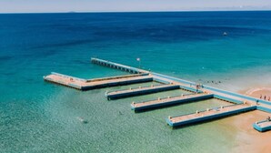 Eine Luftaufnahme zeigt einen Steg, der in ein klares, blaues Meer hinausführt, mit einem Sandstrand am Ende.