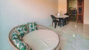 A room with a round wicker sofa with a cushion, a dining table with chairs, and a kitchen area visible through an open doorway. - Casa Refúgio (Cruz)