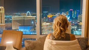 Eine Person sitzt auf einem Sofa mit einer Decke und blickt durch ein großes Fenster auf die Stadt.