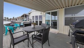 A covered outdoor deck with a round table and chairs, a grill, and a view of the beach and houses in the background. - Little Launch - Waterfront, SpaceX Views, Pools (Port Isabel)