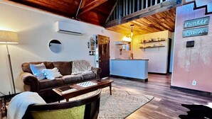 A cozy living room with a brown leather sofa, a wooden coffee table, and a round rug. A kitchen area is visible in the background with a sign reading "Pioneer Cabin." - Pioneer Cabin Retreat (Gasquet)