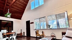 A spacious room with a high ceiling, large windows, a wooden ceiling, and a dining area with chairs and a table. - Pioneer Cabin Retreat (Gasquet)