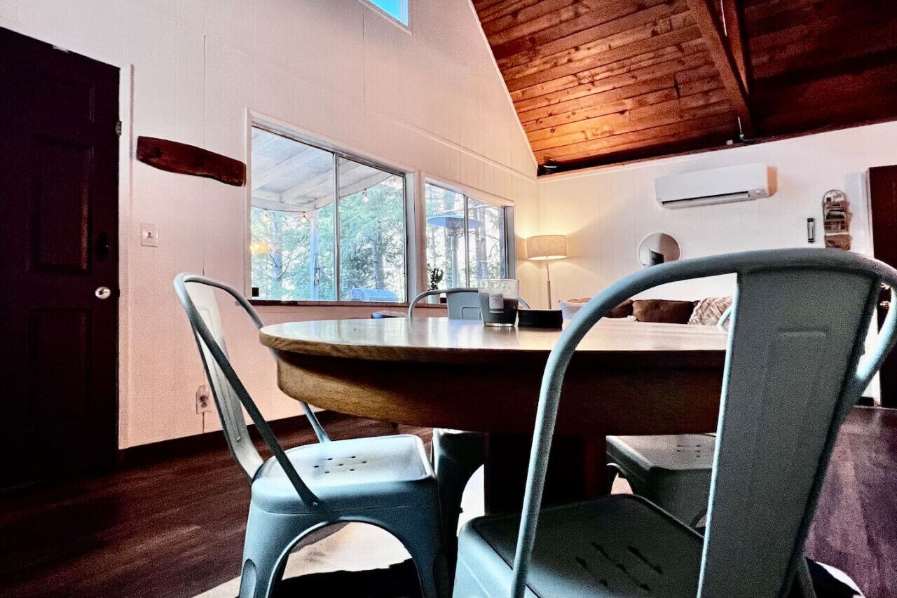 A dining area with a wooden table and metal chairs, large windows, and an air conditioning unit on the wall.