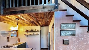 A kitchen with wooden ceiling beams, a built-in island, and a refrigerator. There are signs on the wall reading "Pioneer Cabin" and various handwritten notes and drawings. - Pioneer Cabin Retreat (Gasquet)