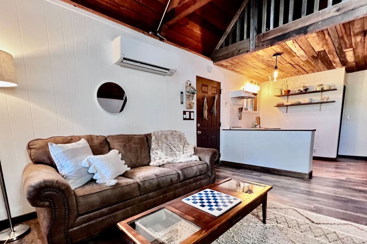 Ein Wohnzimmer mit braunem Ledersofa, einem Couchtisch mit Schachbrett und einer Küchenzeile mit weißen Schränken und Holzbalkendecke.